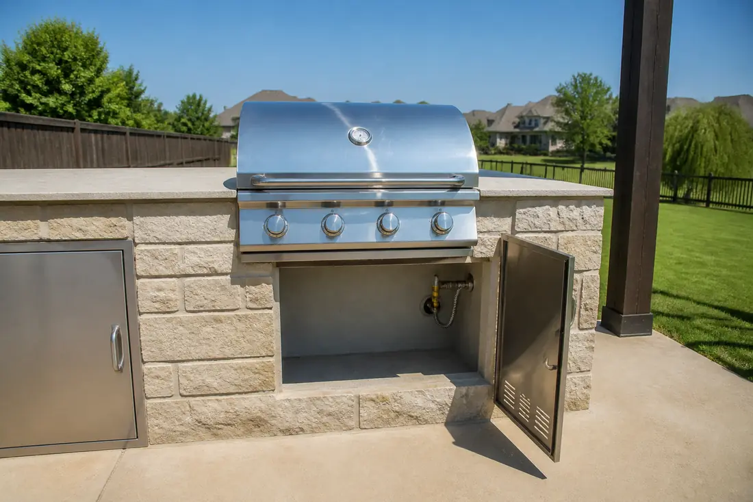 A stone-faced grill island with its access door open, showing the gas shutoff valve and connector inside